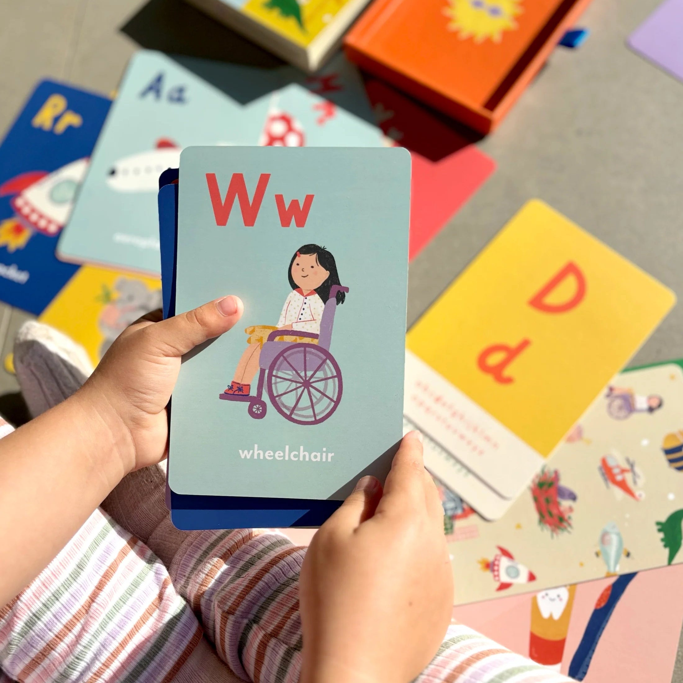 Child holding an alphabet card with a child in a wheelchair, surrounded by other colorful alphabet cards.