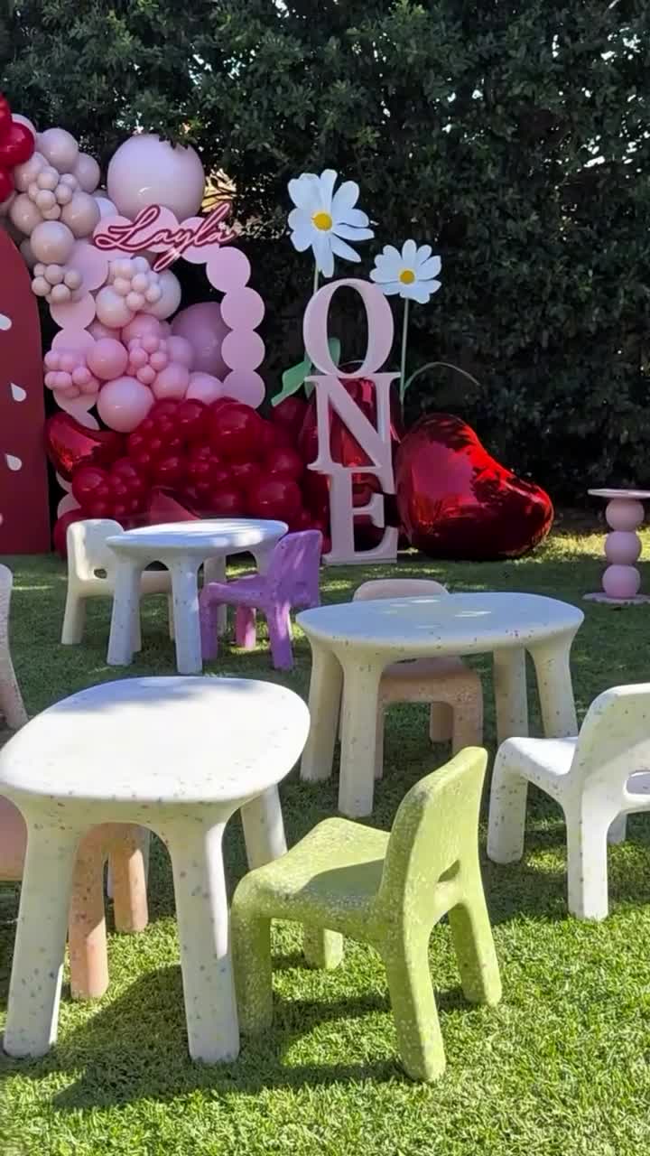 Children's play area with colorful chairs and tables on grass, decorated with balloons and 'ONE' letter set.