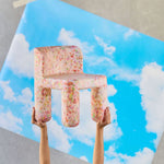 Colorful chair-shaped pillow held by hands against a blue sky with clouds background