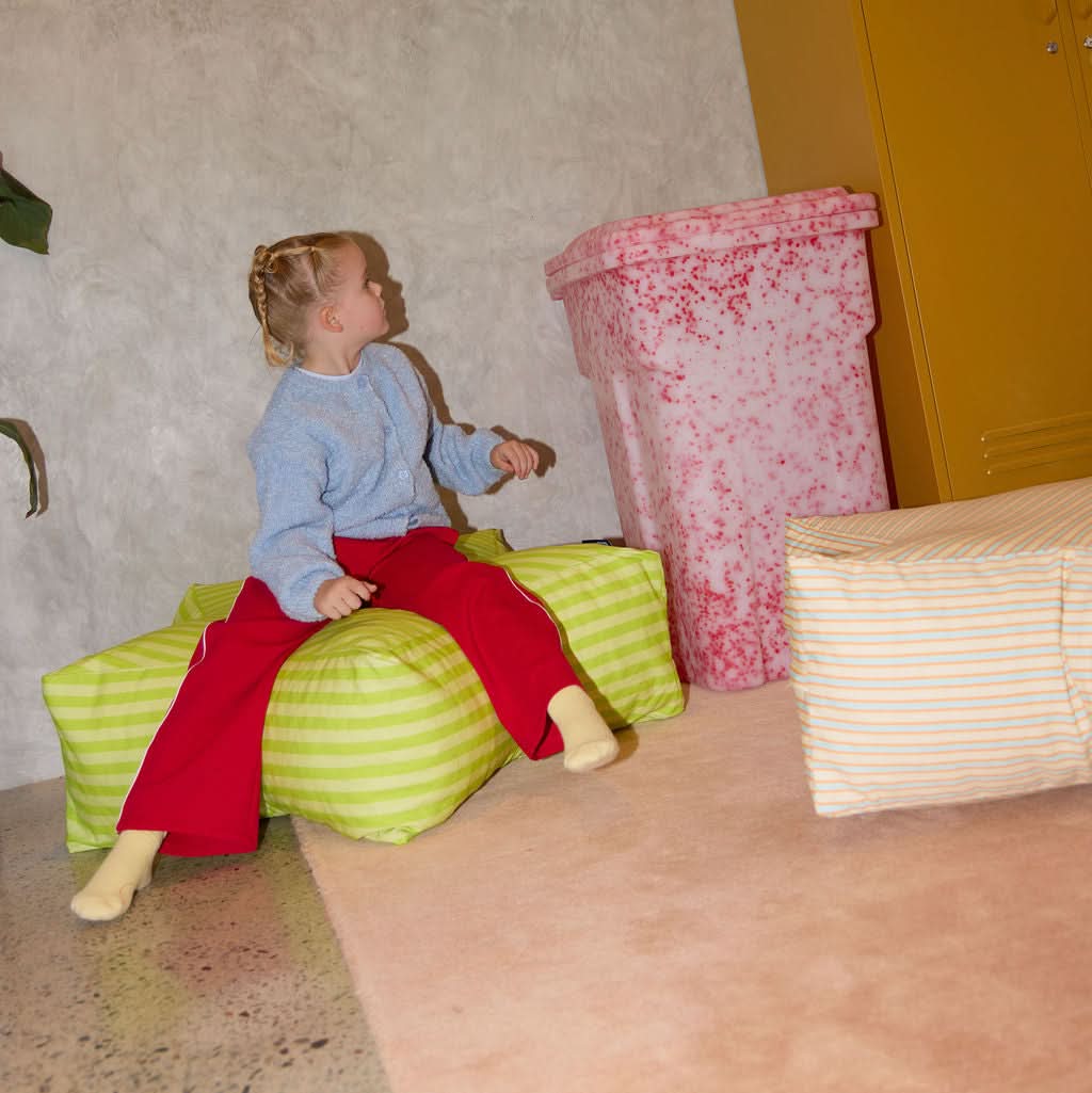 Child sitting on a green striped ottoman in a room with a yellow locker and pink trash can.