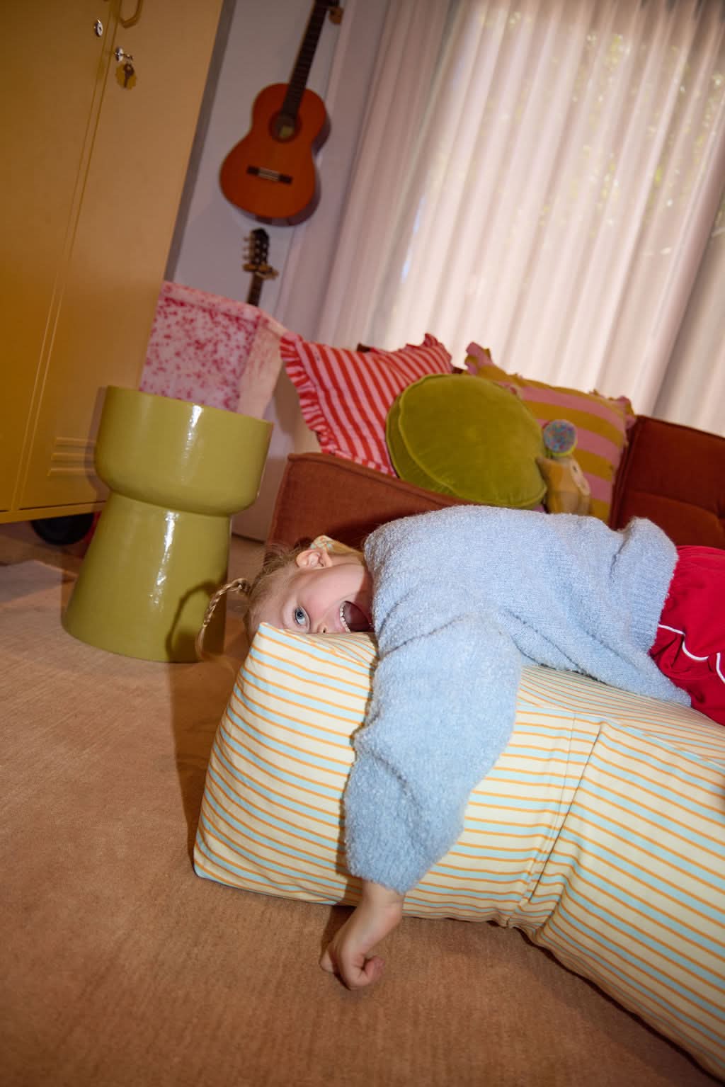 Child in a blue blanket lying on a striped pillow with a guitar and colorful cushions in the background.