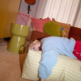 Child in a blue blanket lying on a striped pillow with a guitar and colorful cushions in the background.