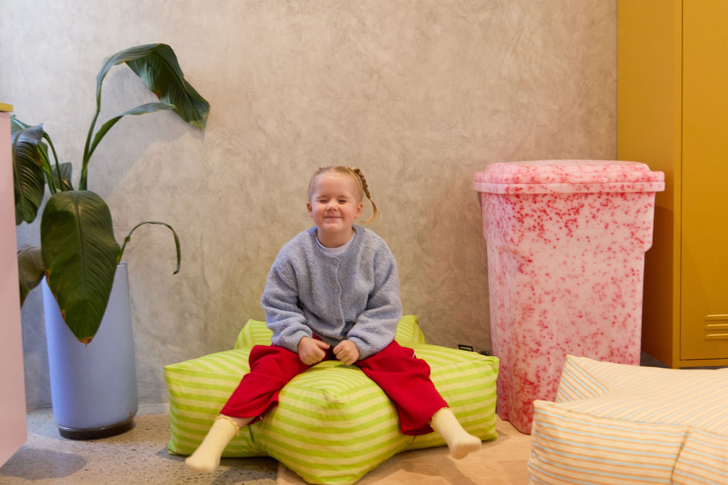 Child sitting on a green bean bag chair in a room with a pink textured wall and plant.