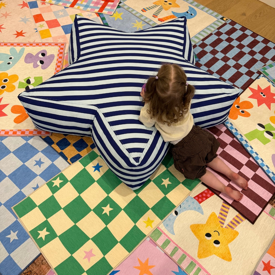 A young child sits on a large star-shaped floor cushion with navy and white stripes, placed on a colorful patchwork quilted play mat.