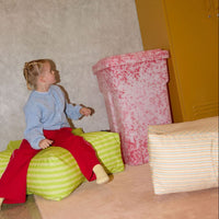 Child sitting on a green striped ottoman in a room with a yellow locker and pink trash can.
