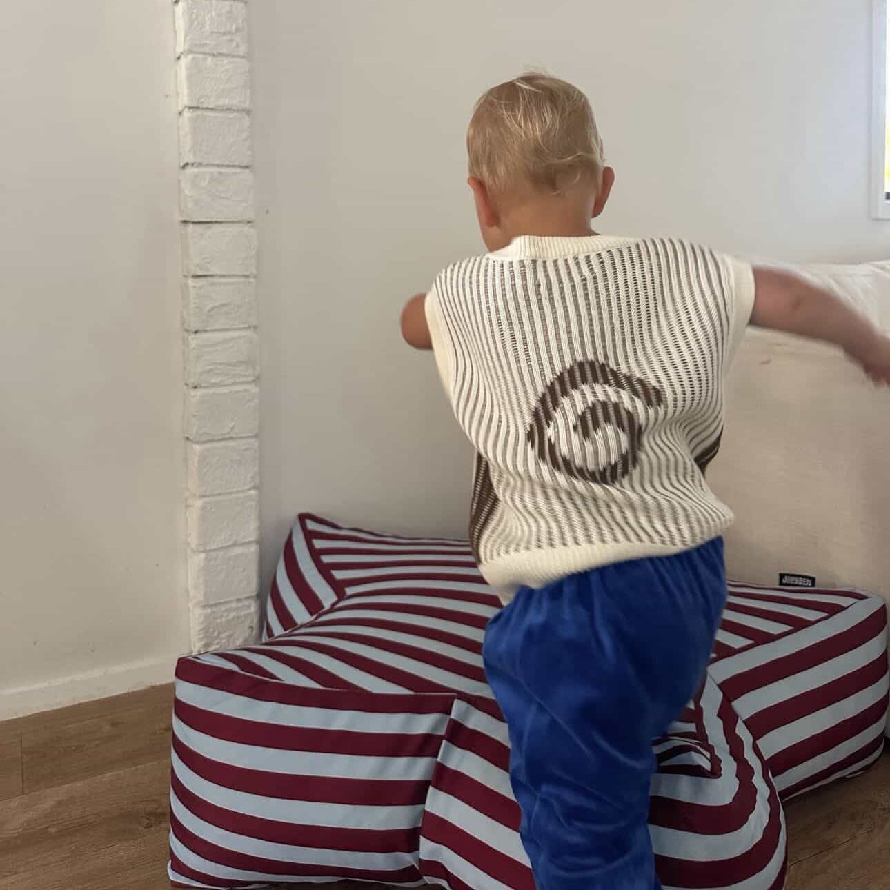 Child sitting on a striped cushion in a room with white walls and a brick column.