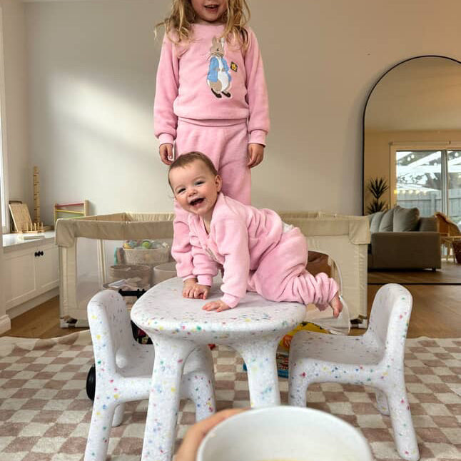Two children in pink outfits playing on a small table in a home setting, with a hand holding a cup of coffee in the foreground.