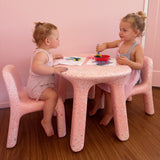 Two young girls sitting at a pink table with chairs in a room with pink walls.