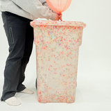 Person holding a pink balloon over a colorful trash can on a white background