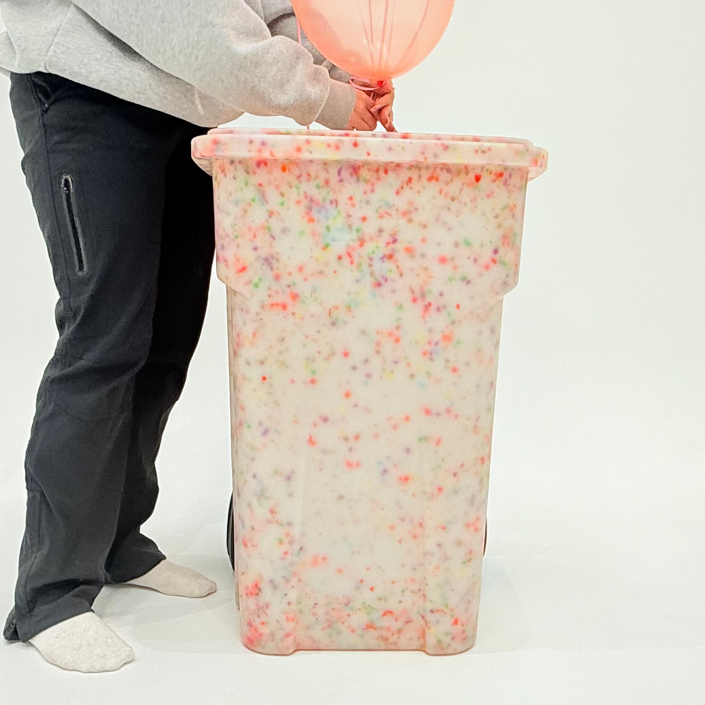 Person holding a pink balloon over a colorful trash can on a white background