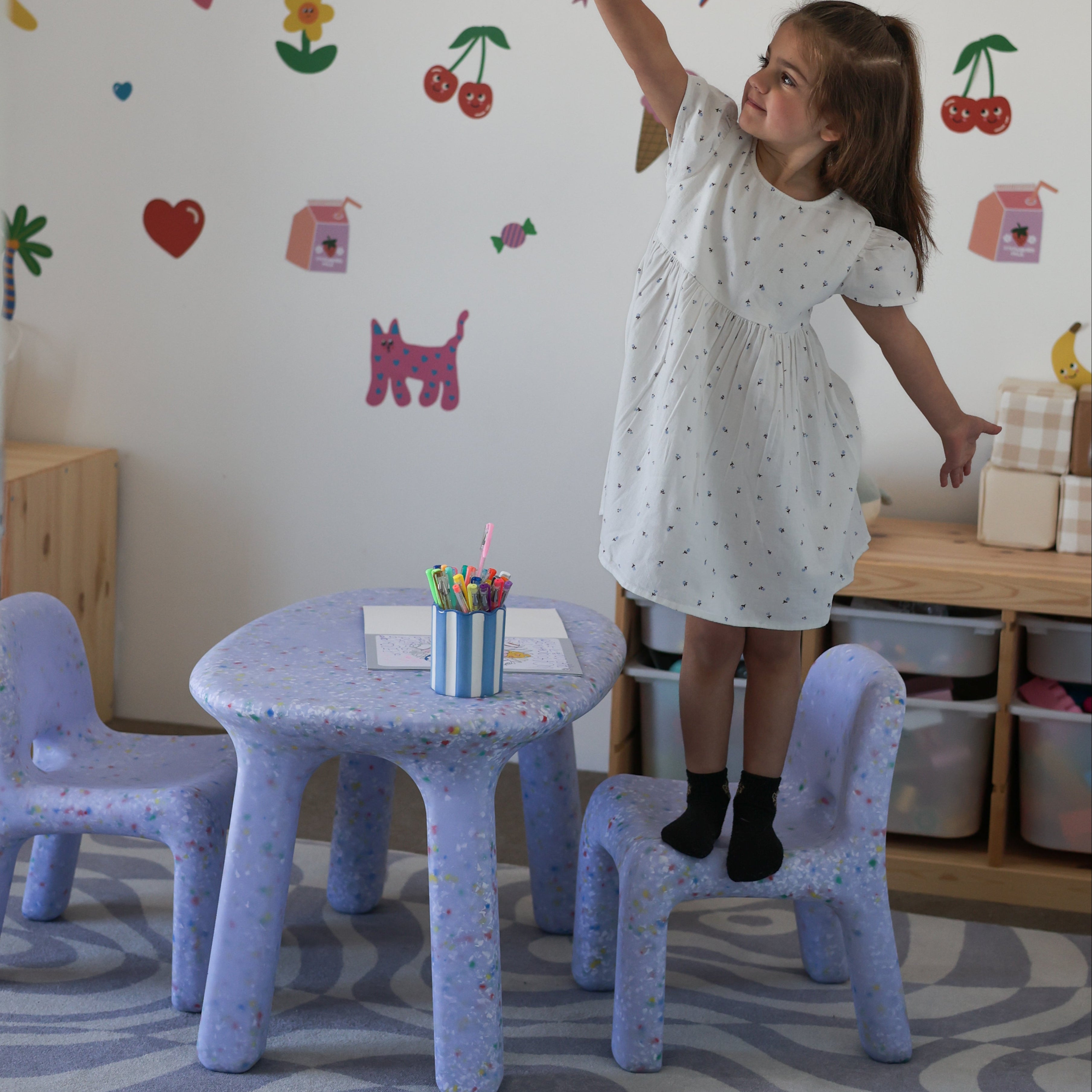 Child in a white dress standing on a chair in a room with colorful wall stickers.