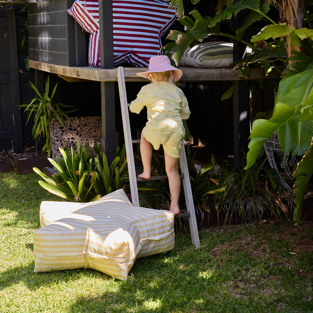 Child climbing a ladder to a playhouse in a garden with striped furniture.