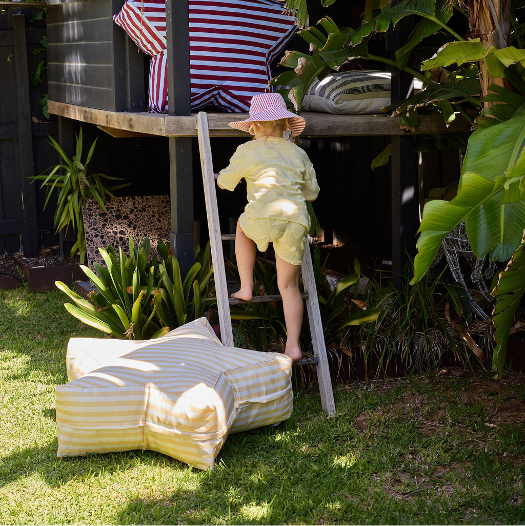 Child climbing a ladder to a playhouse in a garden with striped furniture.