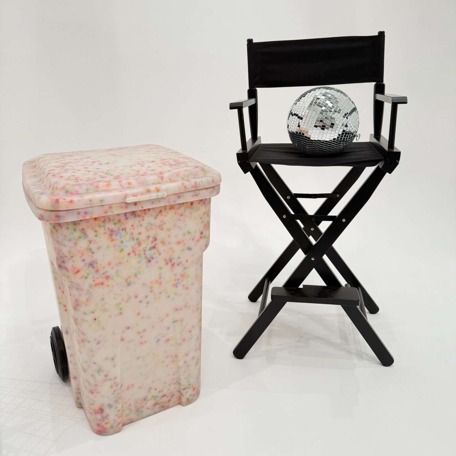Pink patterned trash can next to a black director's chair with a disco ball on a white background