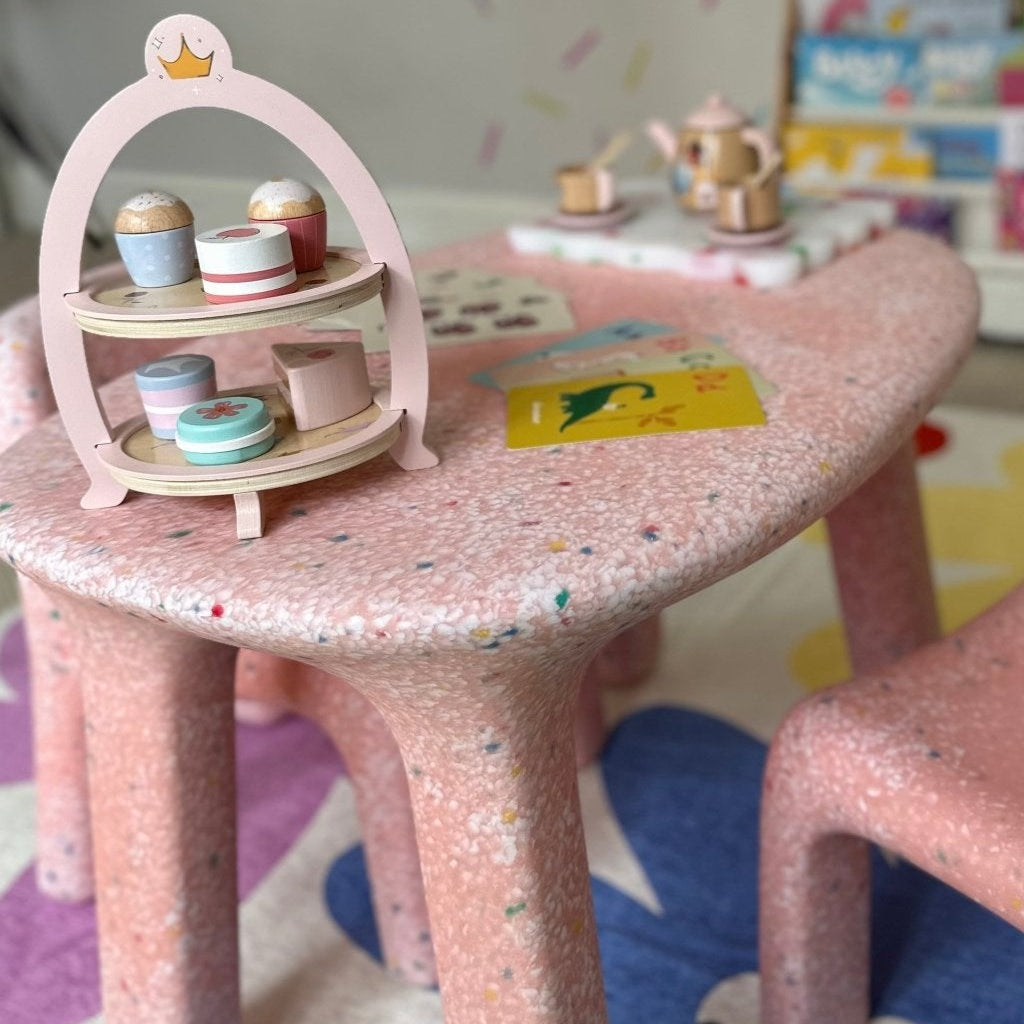 Pastel pink speckled children's table and chair set on a colorful rug with a playful face design. A wooden toy tea set and macaron stand sit on the table. A bookshelf filled with children's books stands against a white wall decorated with multicolored sprinkle-like patterns.