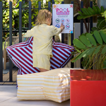 Child in yellow outfit standing on striped cushions next to a pool sign with plants and furniture in the background.